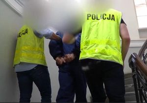 Zdjęcie przedstawia prowadzenie zatrzymanej osoby przez nieumundurowanych policjantów.