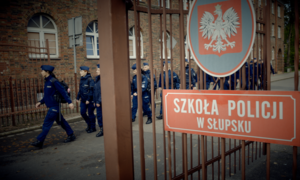 Grupa policjantów maszeruje, na pierwszym planie tabliczka na bramie z napisem Szkoła Policji w Słupsku, a nad nim godło Polski.