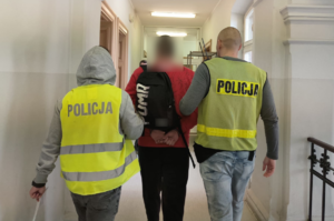 Policjanci ubrani w odblaskowe kamizelki z napisem policja prowadzą zatrzymanego mężczyznę