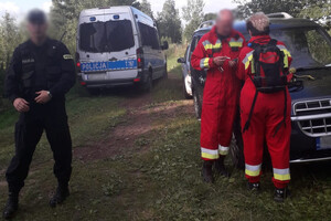 Na zdjęciu widoczny jest policjant wraz ze strażakami , w trakcie akcji poszukiwawczej. W tle znajduje się radiowóz i dwa samochody osobowe.