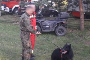 Na zdjęciu widoczny jest żołnierz wraz ze strażakiem i psem tropiącym. W tle znajduje się quad.