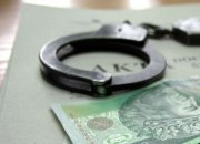 Zdjęcie przedstawia policyjne kajdanki, banknot pieniędzy i teczkę z aktami dochodzenia/śledztwa,