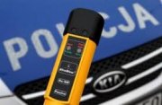 Zdjęcie przedstawia urządzenie do pomiaru stanu trzeźwości. W tle znajduje się policyjny radiowóz.