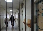 Zdjęcie przedstawia Policyjne Pomieszczenie Dla Osób Zatrzymany. W tle znajdują się dwie osoby.