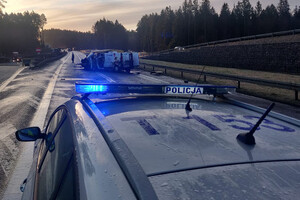 Zdjęcie przedstawia fragment radiowozu policyjnego. W tle znajdują się dwa pojazdy, które uczestniczyły w zdarzeniu drogowym.