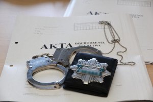 Zdjęcie przedstawia kajdanki policyjne, odznakę i teczkę akt dochodzeniowo-śledczych.