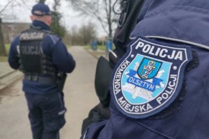 Zdjęcie przedstawia naszywkę KMP Olsztyn a w tle umundurowany policjant.