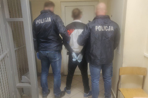 Zdjęcie przedstawia dwóch nieumundurowanych policjantów prowadzących zatrzymanego mężczyznę.