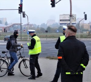 Zdjęcie przedstawia akcję rozdawania odblasków, która miała miejsce w Olsztynie (25.09.2020) w godzinach porannego szczytu komunikacyjnego. Działania przeprowadzono w rejonie skrzyżowania ulicy Leonharda z Al. Piłsudskiego w Olsztynie. Na zdjęciach widoczni policjanci ubrani w kamizelki odblaskowe a w rękach trzymający odblaski, które przekazywane są przechodniom oraz rowerzystom. Na zdjęciu również widoczny jest ksiądz - kapelan Komendy Miejskiej Policji w Olsztynie ks. Arkadisuz Suchowiecki, który ónież aktywnie włączył się w działania mające na celu poprawę bezpieczeństwa niechronionych uczestników ruchu drogowego.