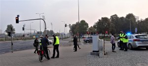 Zdjęcie przedstawia akcję rozdawania odblasków, która miała miejsce w Olsztynie (25.09.2020) w godzinach porannego szczytu komunikacyjnego. Działania przeprowadzono w rejonie skrzyżowania ulicy Leonharda z Al. Piłsudskiego w Olsztynie. Na zdjęciach widoczni policjanci ubrani w kamizelki odblaskowe a w rękach trzymający odblaski, które przekazywane są przechodniom oraz rowerzystom. Na zdjęciu również widoczny jest ksiądz - kapelan Komendy Miejskiej Policji w Olsztynie ks. Arkadisuz Suchowiecki, który ónież aktywnie włączył się w działania mające na celu poprawę bezpieczeństwa niechronionych uczestników ruchu drogowego.