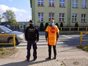 Zdjęcie przedstawia umundurowanego policjanta oraz mężczyznę regulującego ruchem przed przejściem dla pieszych. Na drugim planie znajduje się fragment samochodu, który zatrzymał się przed przejściem dla pieszych. W tle budynek szkoły podstawowej.