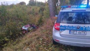Zdjęcie przedstawia miejsce wypadku w pobliżu miejscowości Głotowo. Na pierwszym planie znajduje się fragment policyjnego radiowozu. W tle w przydrożnym rowie znajduje się uszkodzone auto koloru czerwonego.