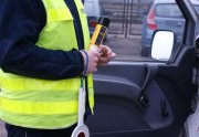 Zdjęcie przedstawia fragment umundurowanego policjanta oraz fragment auta. Policjant trzyma w ręku trzyma urządzenia do badania stanu trzeźwości oraz tarcze do zatrzymania pojazdów.