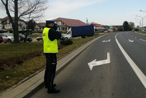 Zdjęcie przedstawia umundurowanego policjanta wykonującego pomiar prędkości. W tle znajduje się jezdnia.
