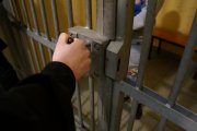 Zdjęcie przedstawia fragment mężczyzny za kratami. Na pierwszym planie znajduje się fragment ręki.