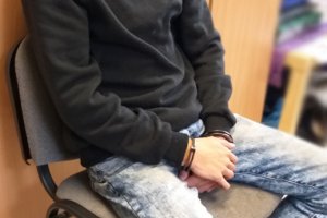 Zdjęcie przedstawia mężczyznę w kajdankach założonych na ręce trzymane z przodu. Mężczyzna siedzi na krześle przy biurku.