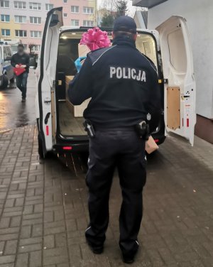 Zdjęcie przedstawia umundurowanego policjanta, który pomaga w zapakowaniu prezentów do białego busa.