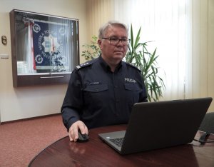 Zdjęcie przedstawia insp. Piotra Zabuskiego siedzącego przed laptopem. W tle znajduje się sztandar KMP w Olsztynie w gablocie.