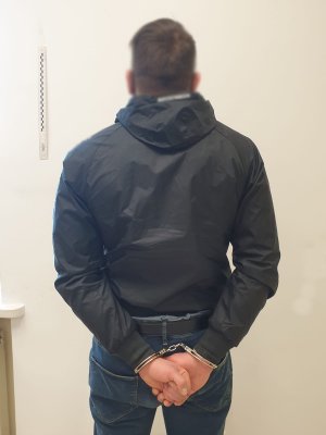 Zdjęcie przedstawia zatrzymanego mężczyznę stojącego przed ścianą. Mężczyzna ma założone kajdanki na ręce trzymane z tyłu.