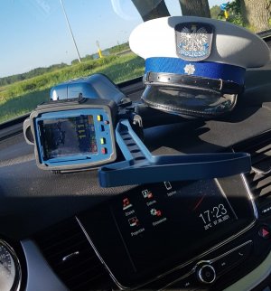 Ilustracja przedstawia policjant oraz urządzenie służące do pomiaru prędkości. Urządzenie posiada możliwość rejestrowania obrazu w związku z czym na jednym ze zdjęć widoczna jest prędkość 11 km/h.