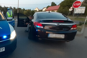 Zdjęcie przedstawia samochód i siedzącego w nim mężczyznę. Obok fragment policyjnego radiowozu oraz policjant.