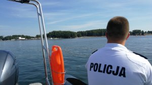 Zdjęcie przedstawia fragment policjanta na łodzi a w tle jezioro.