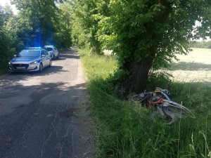 Zdjęcie przedstawia miejsce zdarzenia drogowego na trasie Kamionka-Rozogi. Na pierwszym planie znajduje się motocykl leżący przy drzewie a w tle policyjne dwa radiowozy.