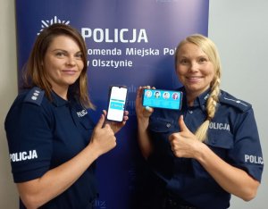 Zdjęcie przedstawia umundurowane policjantki trzymające w ręku telefony a w tle baner z napisem &quot;Komenda Miejska Policji w Olsztynie&quot;.