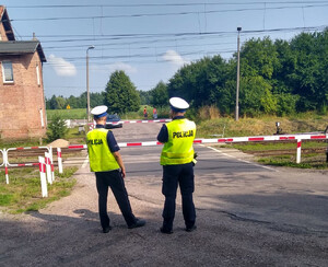 Zdjęcie przedstawia dwóch umundurowanych policjantów stojących przed przejazdem kolejowym.
