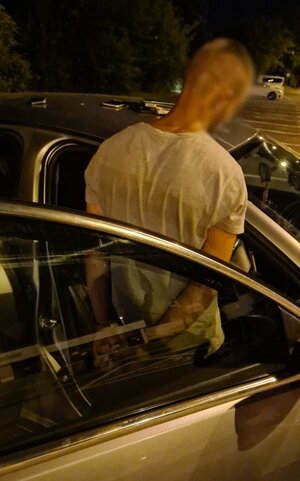 Zdjęcie przedstawia zatrzymanego mężczyznę stojącego przed autem na parkingu.