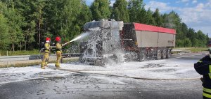 Miejsce pożaru auta ciężarowego wraz z naczepą. Na miejscu widać płonący pojazd ciężarowy wraz z naczepą typu wanna. Zdarzenie ma miejsce na jezdni dwupasmowej. Na zdjęciu widać dym wydobywający się z płonącego pojazdu oraz strażaków gaszących płomienie przy wykorzystaniu węża strażackiego.