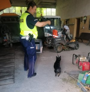 Policjant odprowadzający psa do schroniska. Zdjęcie przedstawia wnętrze garażu. Funkcjonariusz ubrany w kamizelkę odblaskową z napisem POLICJA. Policjant trzyma na smyczy psa maści czarnej.