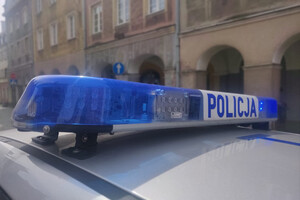 Zdjęcie przestawia fragment policyjnego radiowozu a w tle budynek.