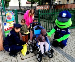 Zdjęcie przedstawia umundurowaną policjantkę, obok maskotkę sierżanta żabusia oraz dzieci o osoba dorosła. W tle budynek szkoły.