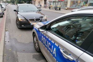 Zdjęcie przedstawia fragment policyjnego radiowozu a obok pojazd marki BMW koloru czarnego. W tle ulica.