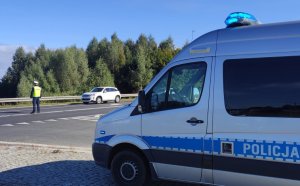 Zdjęcie przedstawia policyjny radiowóz policyjny z napisem POLICJA na boku pojazdu. Przed radiowozem znajduje się jezdnia a na niej stoi policjant regulujący ręcznie ruchem.