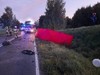 Zdjęcie przedstawia miejsce wypadku drogowego ze skutkiem śmiertelnym. Na zdjęciu widać drogę a na niej porozrzucane elementy uszkodzonych aut. Na zdjęciu widać rozstawiony parawan w kolorze czerwonym.