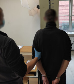 Zdjęcie przedstawia zatrzymanego mężczyznę w pomieszczeniu a obok nieumundurowany policjant.