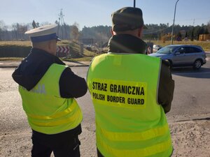 Zdjęcie przedstawia umundurowanego policjanta oraz strażnika granicznego kierującego ruchem drogowym.