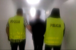 Zdjęcie przedstawia dwóch policjantów prowadzących zatrzymanego mężczyznę.