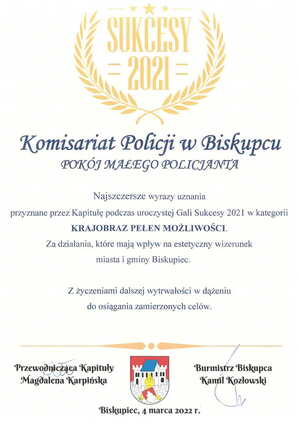 Zdjęcie przedstawia napisy: Komisariat Policji w Biskupcu. Pokój małego policjanta. Najszczersze wyrazy uznania przyznane przez Kapitułę podczas uroczystej Gali Sukcesy 2021 w kategorii Krajobraz Pełen Możliwości. Za działania, które mają wpływ na estetyczny wizerunek miasta i gminy Biskupiec. Z życzenia dalszej wytrwałości w dążeniu do osiągnięcia zamierzonych celów. 
Przewodniczący Kapituły Magdalena Karpińska
Burmistrz Biskupca Kamil Kozłowski 
Biskupiec 4 marca 2022 roku