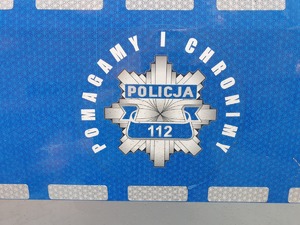 Zdjęcie przedstawia fragment radiowozu z napisem &quot;Policja - Pomagamy i Chronimy&quot;.
