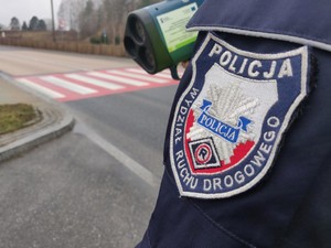 Zdjęcie przedstawia framgment policjanta wraz z miernikiem prędkości w pobliżu oznakowanego przejścia dla pieszych.