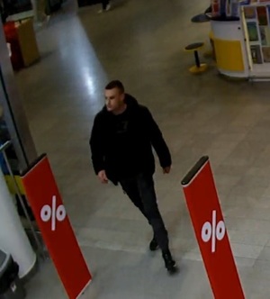 Zdjęcie przedstawia mężczyznę na terenie galerii handlowej odpowiedzialnego za dokonanie kradzieży. Mężczyzna ubrany jest w ciemnych kolorach, jest wysoki i ma ciemne włosy.