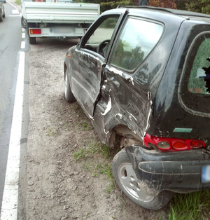 Zdjęcie przedstawia uszkodzony samochód osobowy m-ki Fiat Seicento. Auto w kolorze czarnym ma rozbity tylny lewy bok.