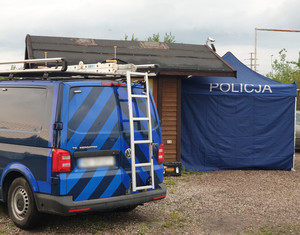 Zdjęcie przedstawia miejsce zdarzenia: ambulans techniki kryminalistycznej oraz policyjny parawan i budynek.