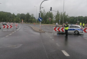 Zdjęcie przedstawia zabezpieczenie zawodów: policjanta wraz z oznakowanym radiowozem. W tle osoby biegnące ulicą.