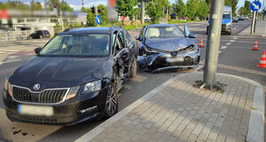Zdjęcie przedstawia auta uczestniczące w kolizji na drodze. W tle budynek oraz policyjny radiowóz.