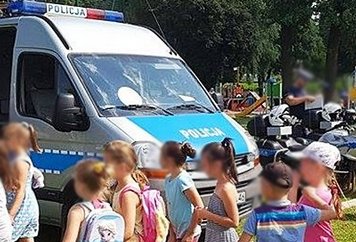 Zdjęcie przedstawia grupę dzieci a w tle policyjny radiowóz oraz motocykl a obok policjant.
