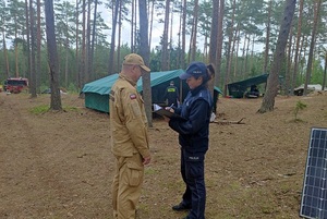 Zdjęcie wykonano na terenie obozowiska leśnego w którym wypoczywa młodzież. Na zdjęciu widoczny funkcjonariusz Policji i Straży Pożarnej a także organizatorzy obozu harcerskiego.